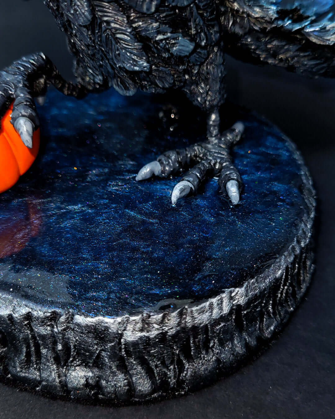 Sculpture d'un corbeau noir irisé bleu, ailes déployées, gardant une citrouille sous ses serres.