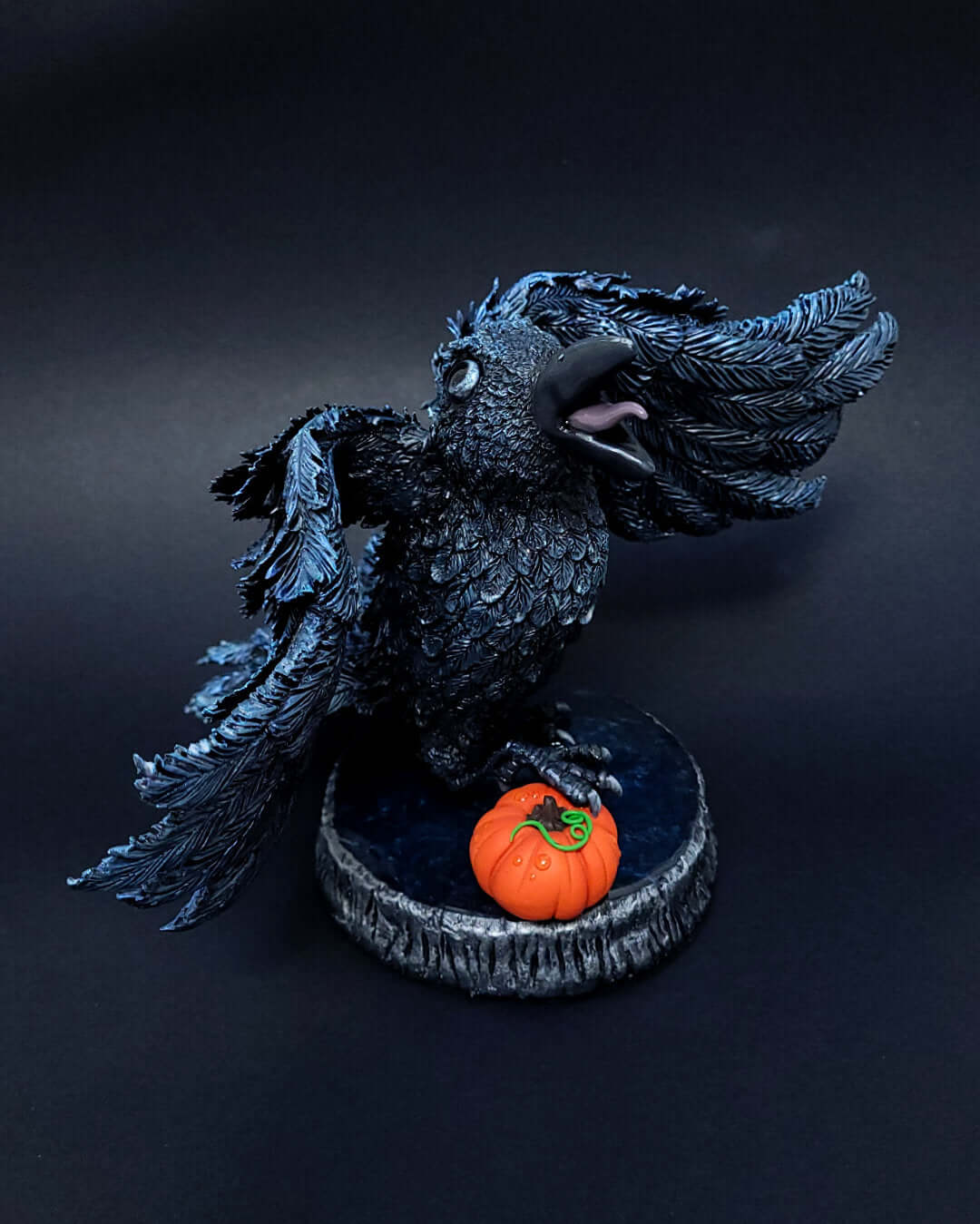 Sculpture d'un corbeau noir irisé bleu, ailes déployées, gardant une citrouille sous ses serres.