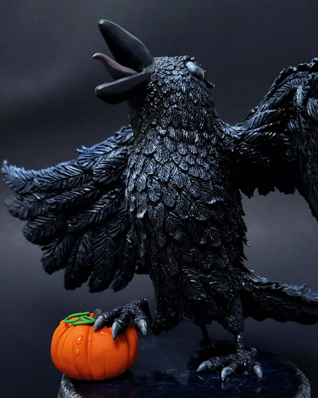Sculpture d'un corbeau noir irisé bleu, ailes déployées, gardant une citrouille sous ses serres.