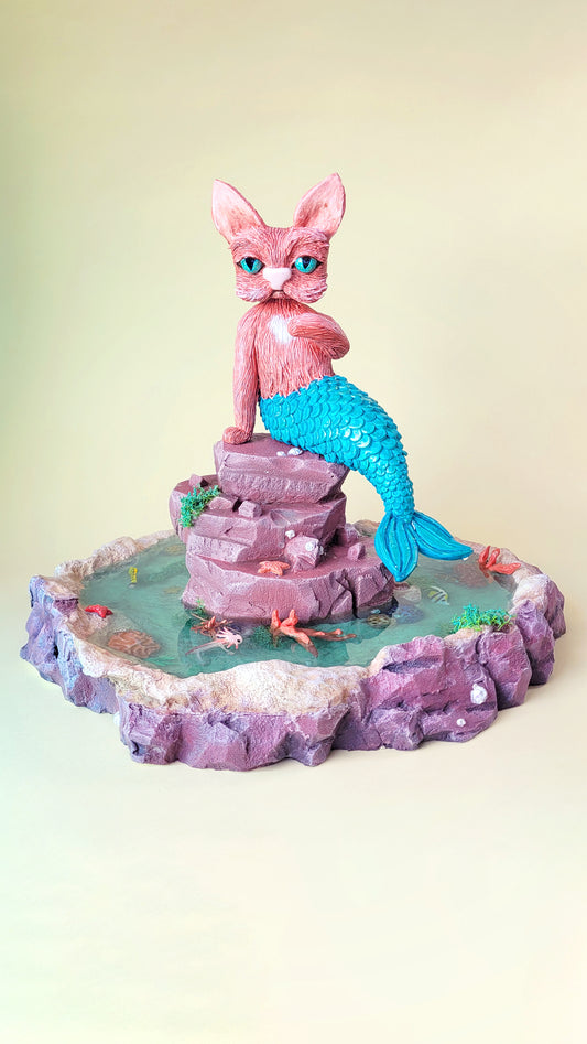 Diorama aquatique avec un gros chat sirène rose et turquoise au centre sur un rocher, entouré de fausse eau et de flore et faune tropicale