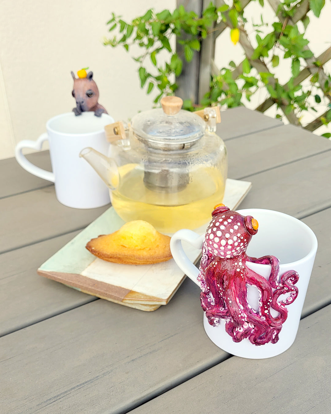 2 tasses de thé, accompagnées d'une théière et de madeleines, décorées de 2 mini figurines en forme de poulpe et de capybara