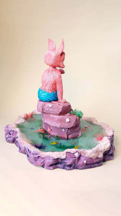 Diorama aquatique avec un gros chat sirène rose et turquoise au centre sur un rocher, entouré de fausse eau et de flore et faune tropicale