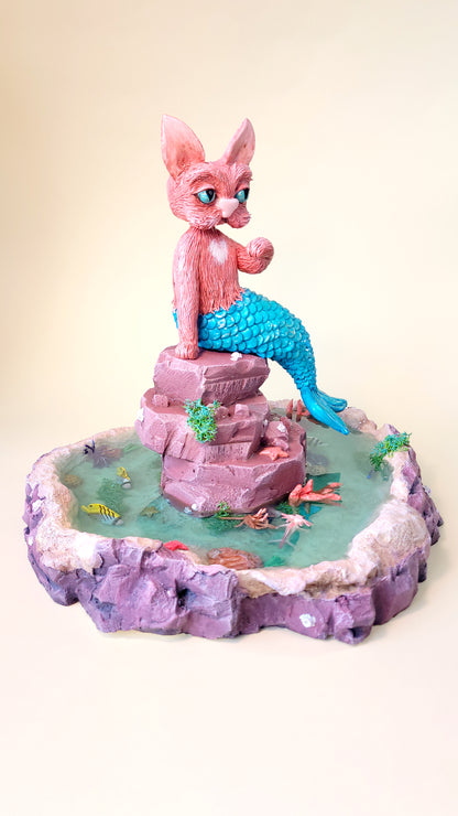 Diorama aquatique avec un gros chat sirène rose et turquoise au centre sur un rocher, entouré de fausse eau et de flore et faune tropicale