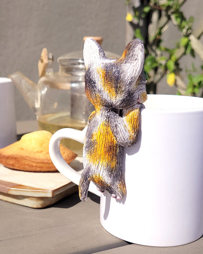 Mini figurine en forme de chat calico accrochée à une tasse de thé vert, accompagné de madeleines