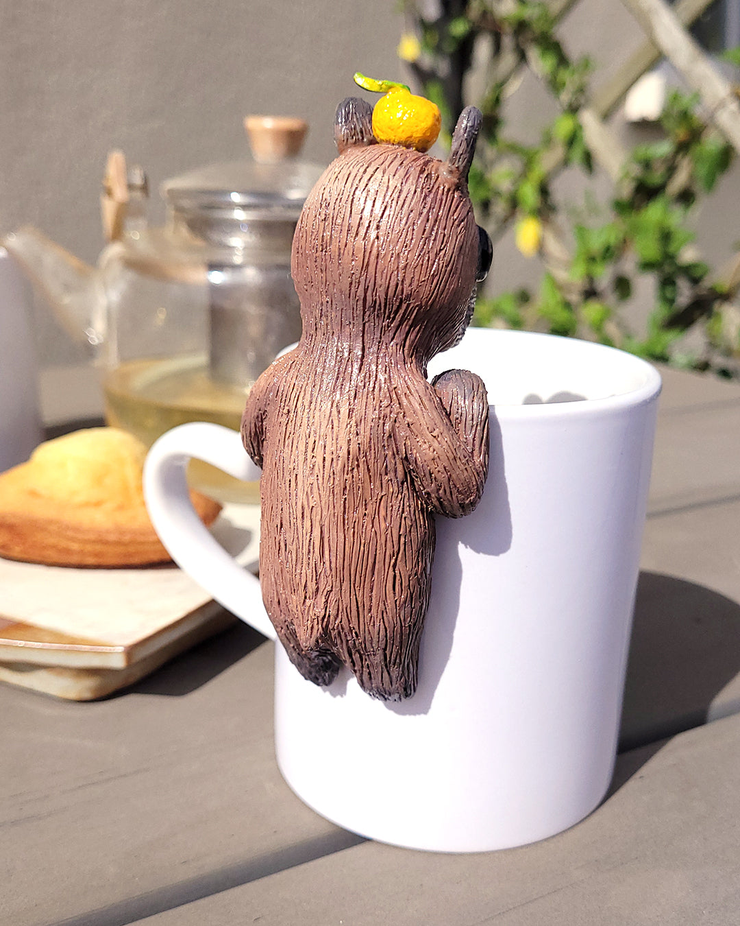 Mini figurine en forme de capybara avec un yuzu sur la tête accrochée à une tasse de thé vert, accompagné de madeleines