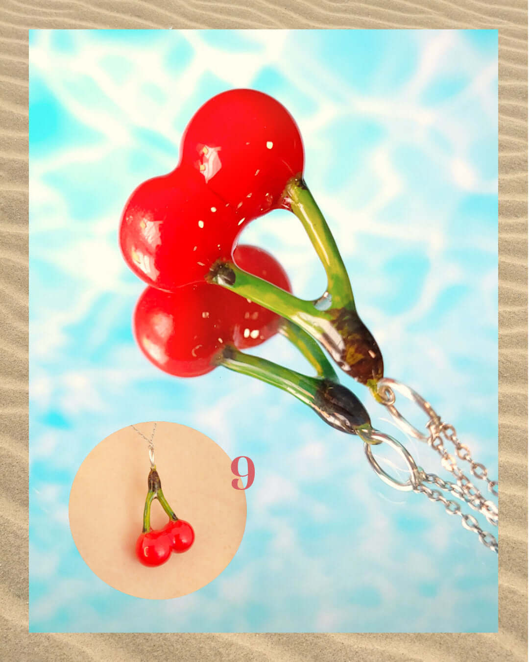 Pendentif rouge et vert en forme de cerises, sur un fond aquatique faisant penser à la surface d'une piscine et encadré de sable.