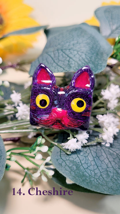 Pins en forme de tête de chat violet et fuchsia aux yeux jaunes qui louchent inspiré de Cheshire, au milieu de fleurs et feuillages