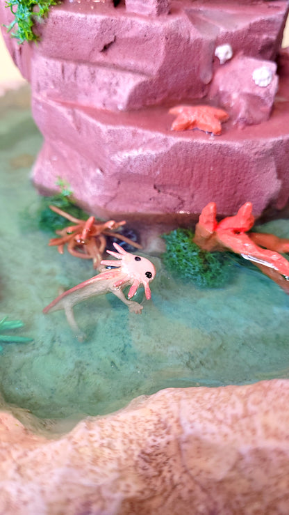 Diorama aquatique avec un gros chat sirène rose et turquoise au centre sur un rocher, entouré de fausse eau et de flore et faune tropicale