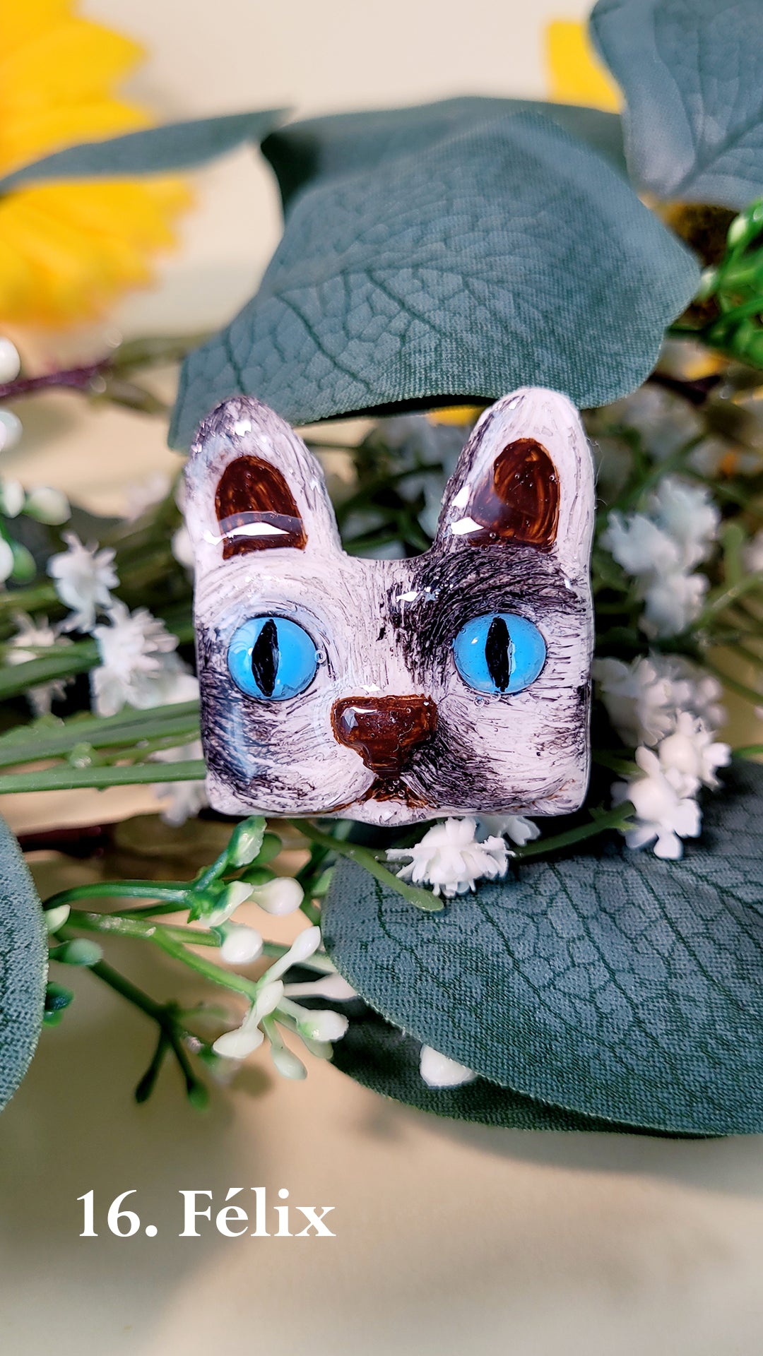 Pins en forme de tête de chat noir et blanc aux yeux bleus, au milieu de fleurs et feuillages