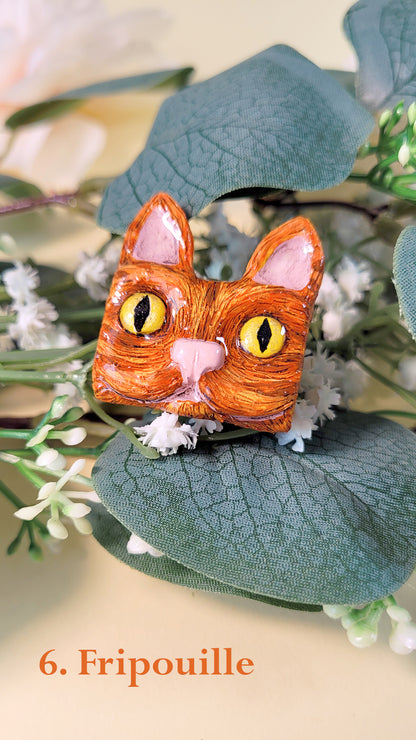 Pins en forme de tête de chat roux aux yeux dorés, au milieu de fleurs et feuillages