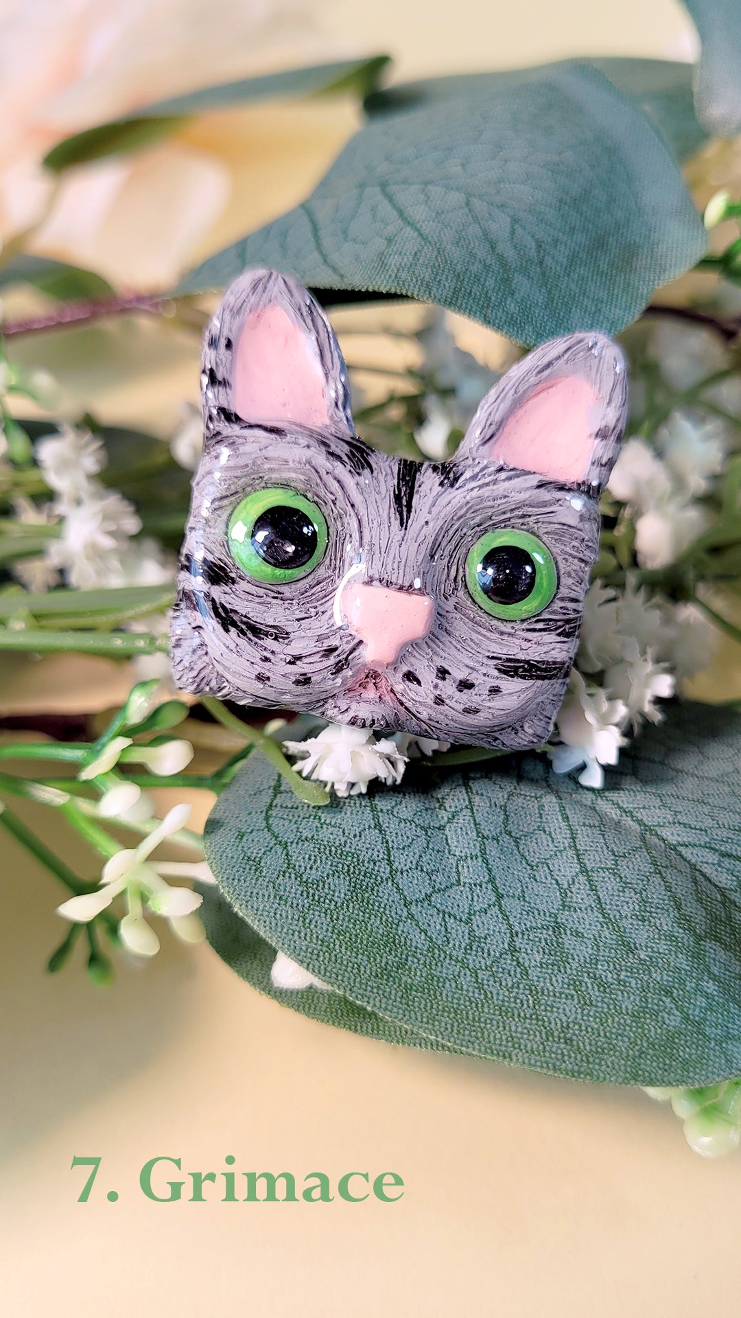 Pins en forme de tête de chat gris rayé aux yeux verts qui louche, au milieu de fleurs et feuillages