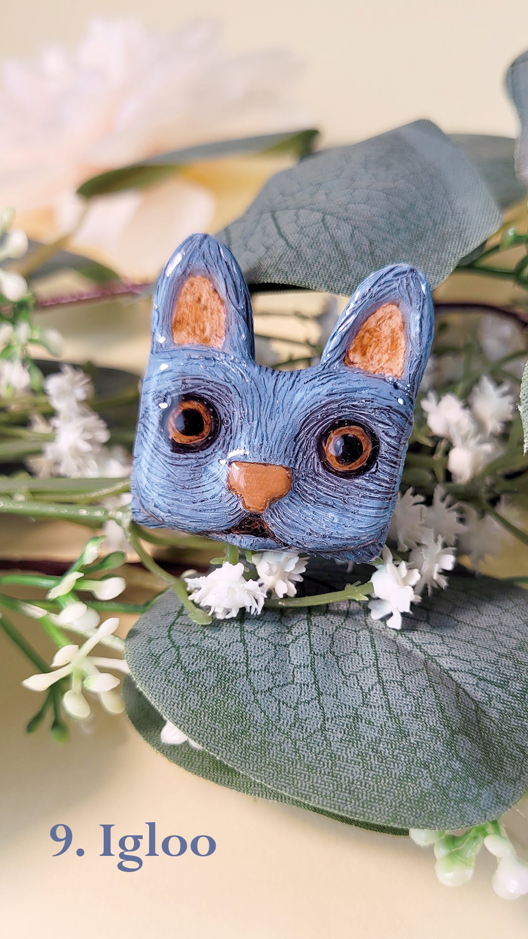 Pins en forme de tête de chat bleu gris aux yeux marrons, au milieu de fleurs et feuillages