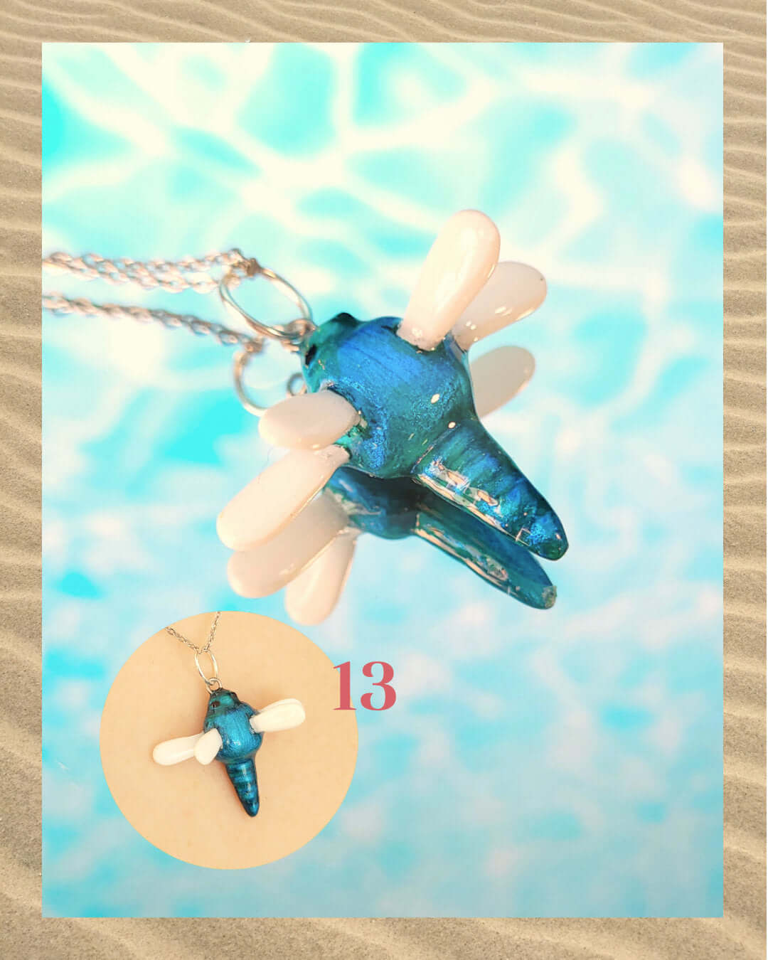 Pendentif irisé bleu en forme de libellule, sur un fond aquatique faisant penser à la surface d'une piscine et encadré de sable.