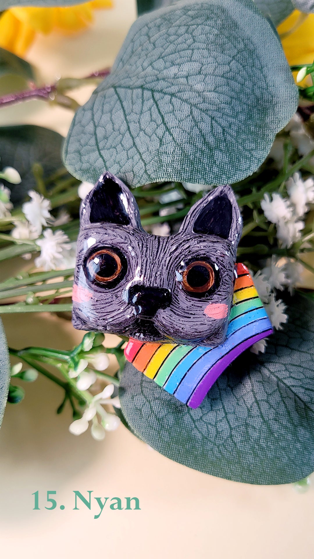 Pins en forme de tête de chat gris aux yeux marrons avec un arc-en-ciel inspiré du Nyan Cat, au milieu de fleurs et feuillages