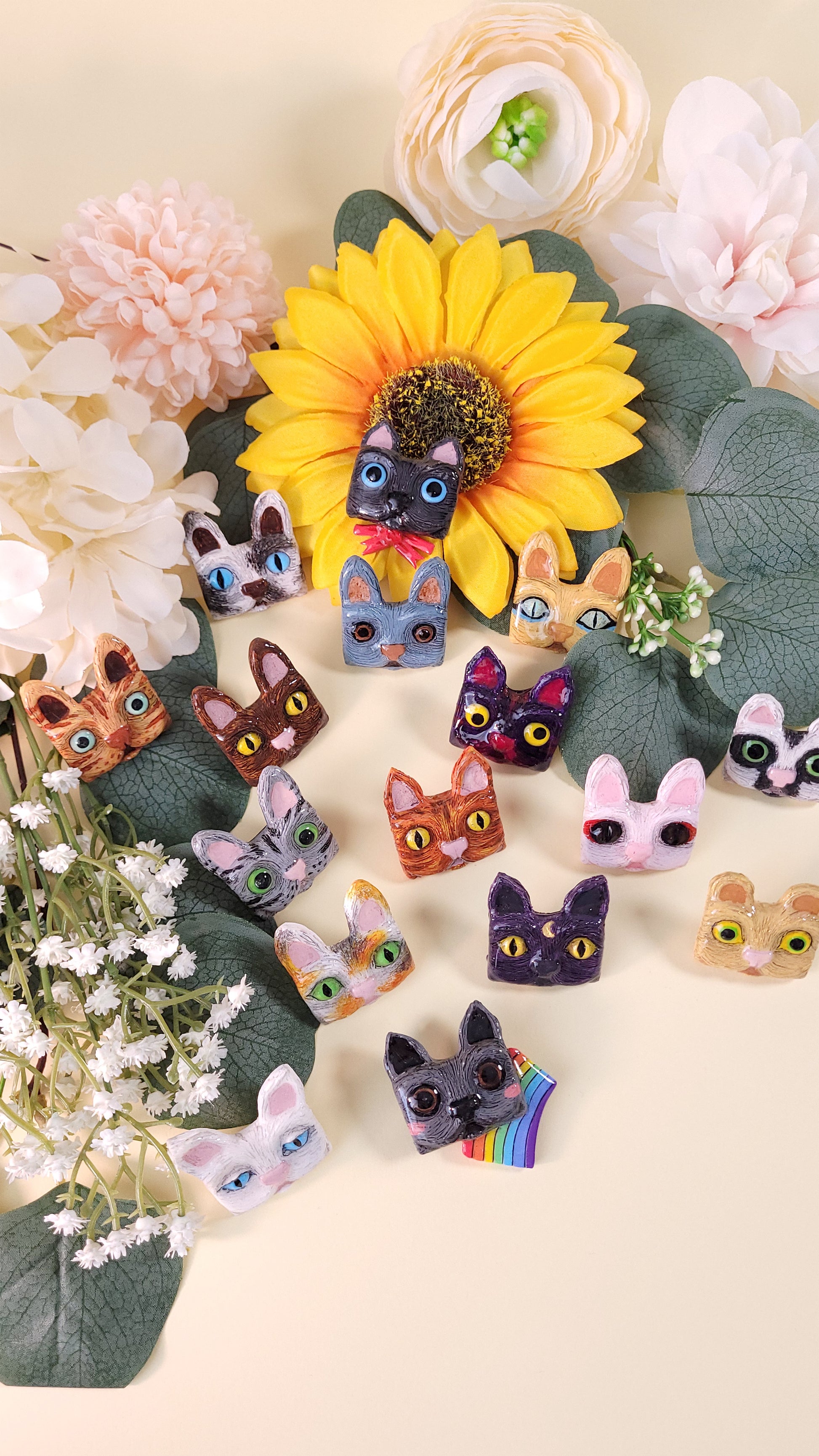 Collection de 16 pins en forme de tête de chat colorées disposés au milieu de fleurs et feuillages