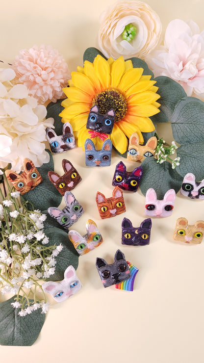 Collection de 16 pins en forme de tête de chat colorées disposés au milieu de fleurs et feuillages