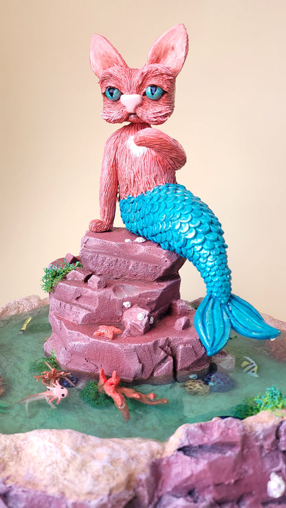 Diorama aquatique avec un gros chat sirène rose et turquoise au centre sur un rocher, entouré de fausse eau et de flore et faune tropicale