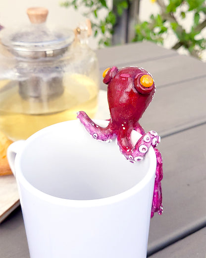 Mini figurine en forme de poulpe violet aux yeux oranges accrochée à une tasse de thé vert