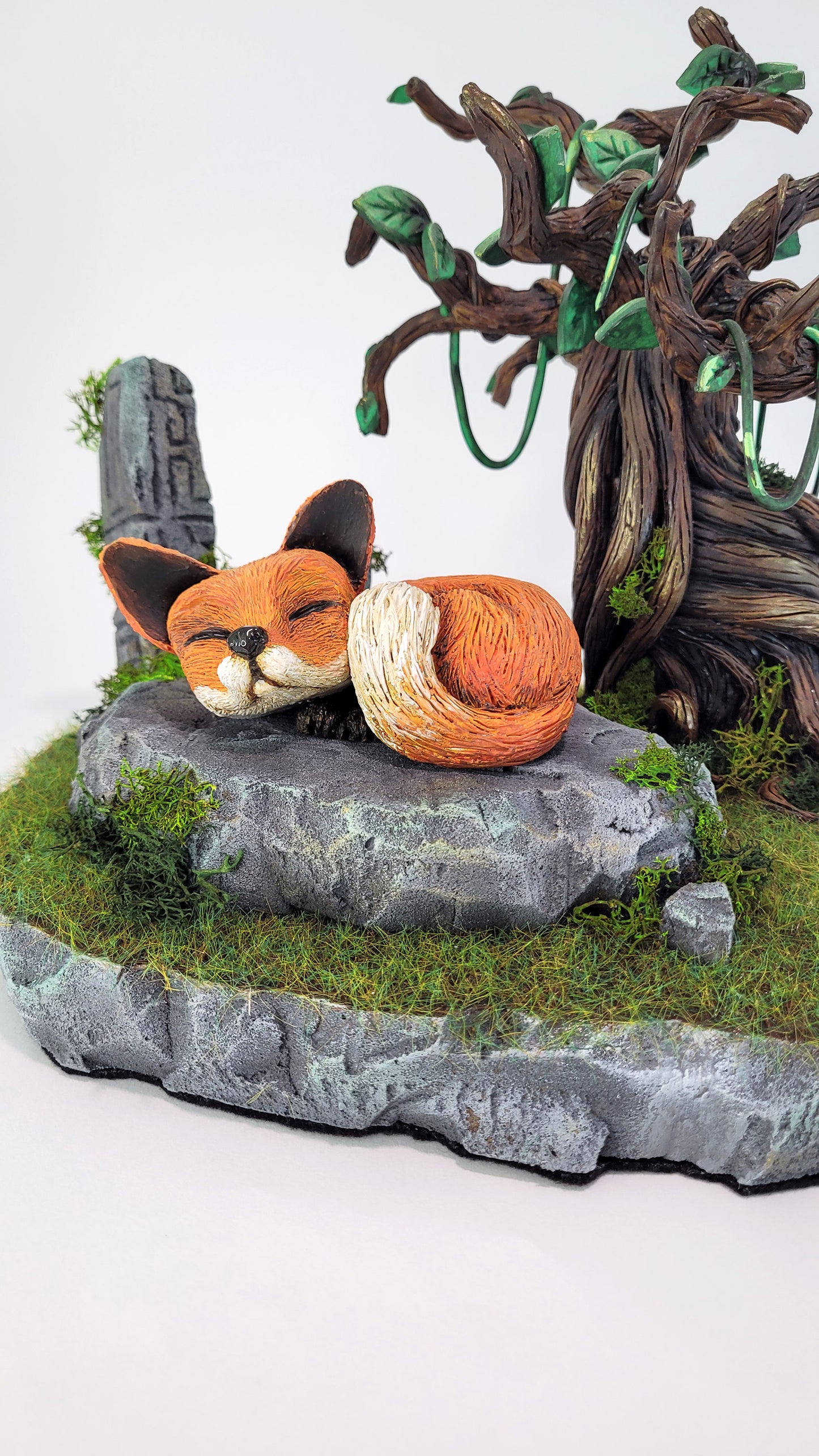 Diorama mettant en scène une figurine de renard dormant sur une pierre à l'ombre d'un arbre sinueux, au milieu de la nature et de ruines abandonnées