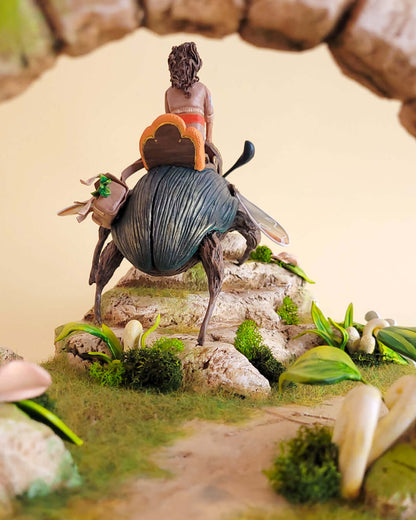 Diorama mettant en valeur une sculpture de machine scarabée géante montée par un personnage féminin. Le décors est riche en verdure.