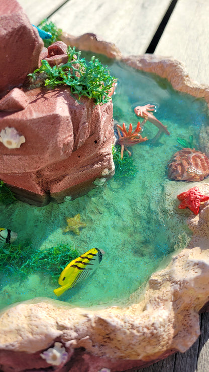 Diorama aquatique avec un gros chat sirène rose et turquoise au centre sur un rocher, entouré de fausse eau et de flore et faune tropicale