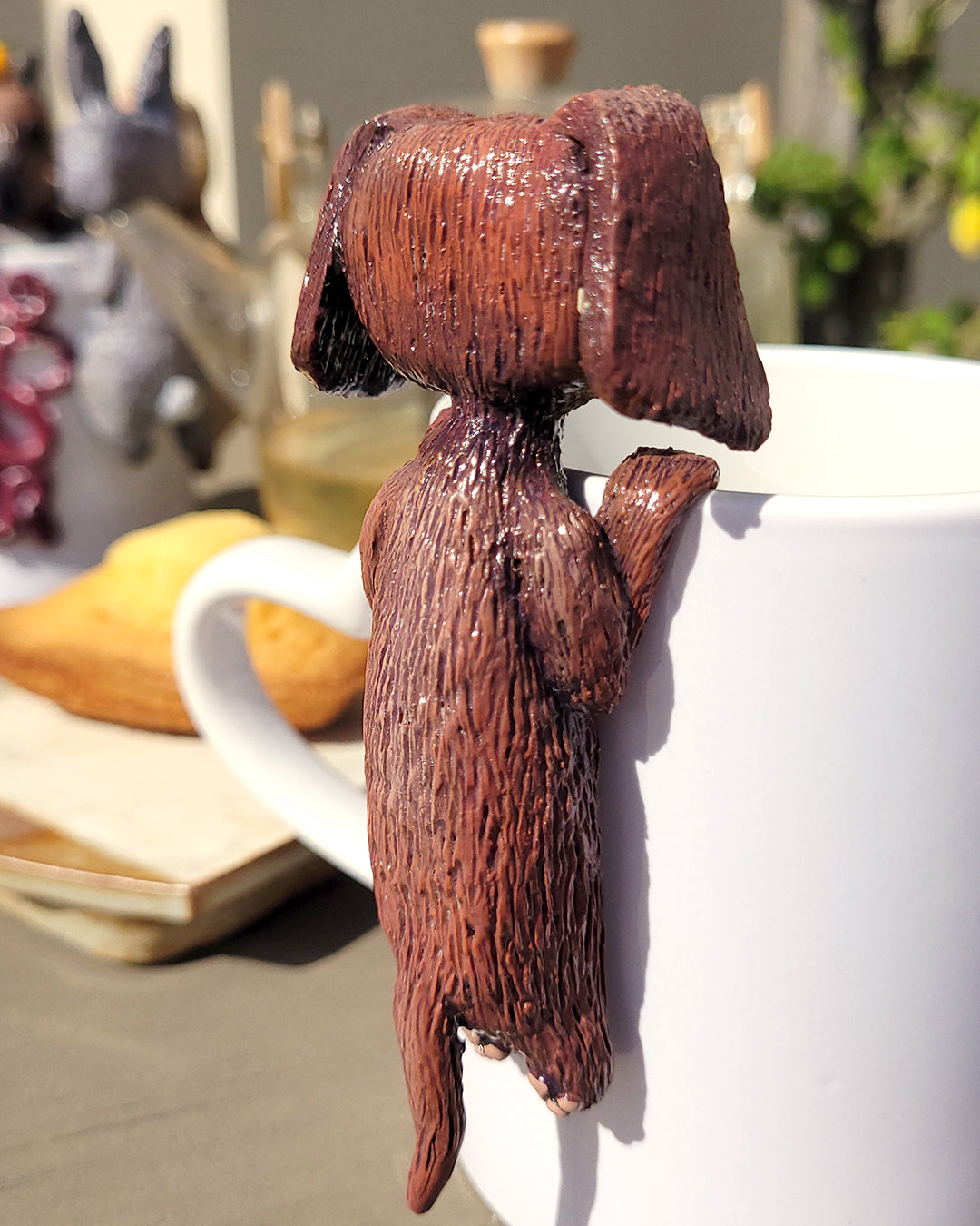 Mini figurine en forme de teckel accrochée à une tasse de thé vert, accompagné de madeleines